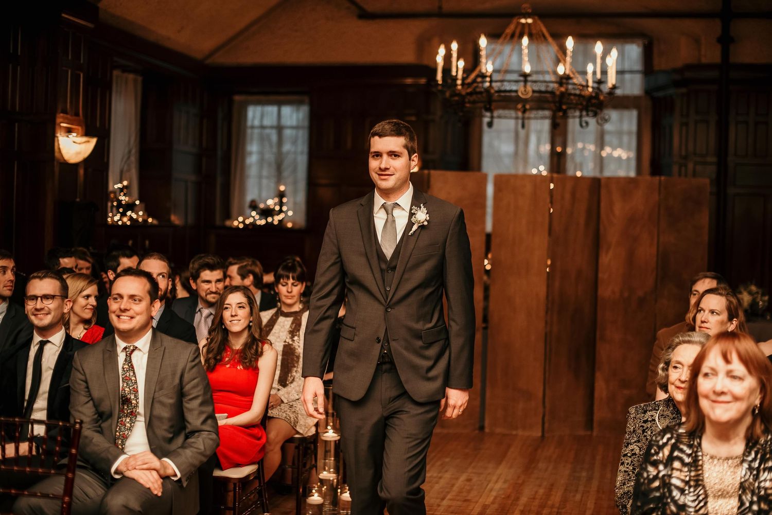 Groom walking down the aisle in a dark suit at an elegant wood-paneled wedding venue with guests seated on both sides.