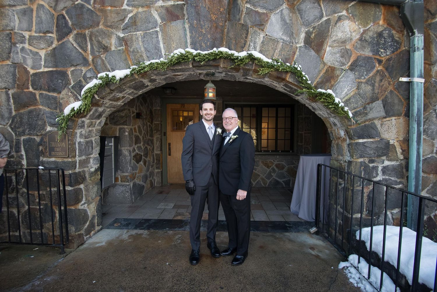 Two men in suits pose under a snow-dusted stone arch with greenery at a winter wedding venue.