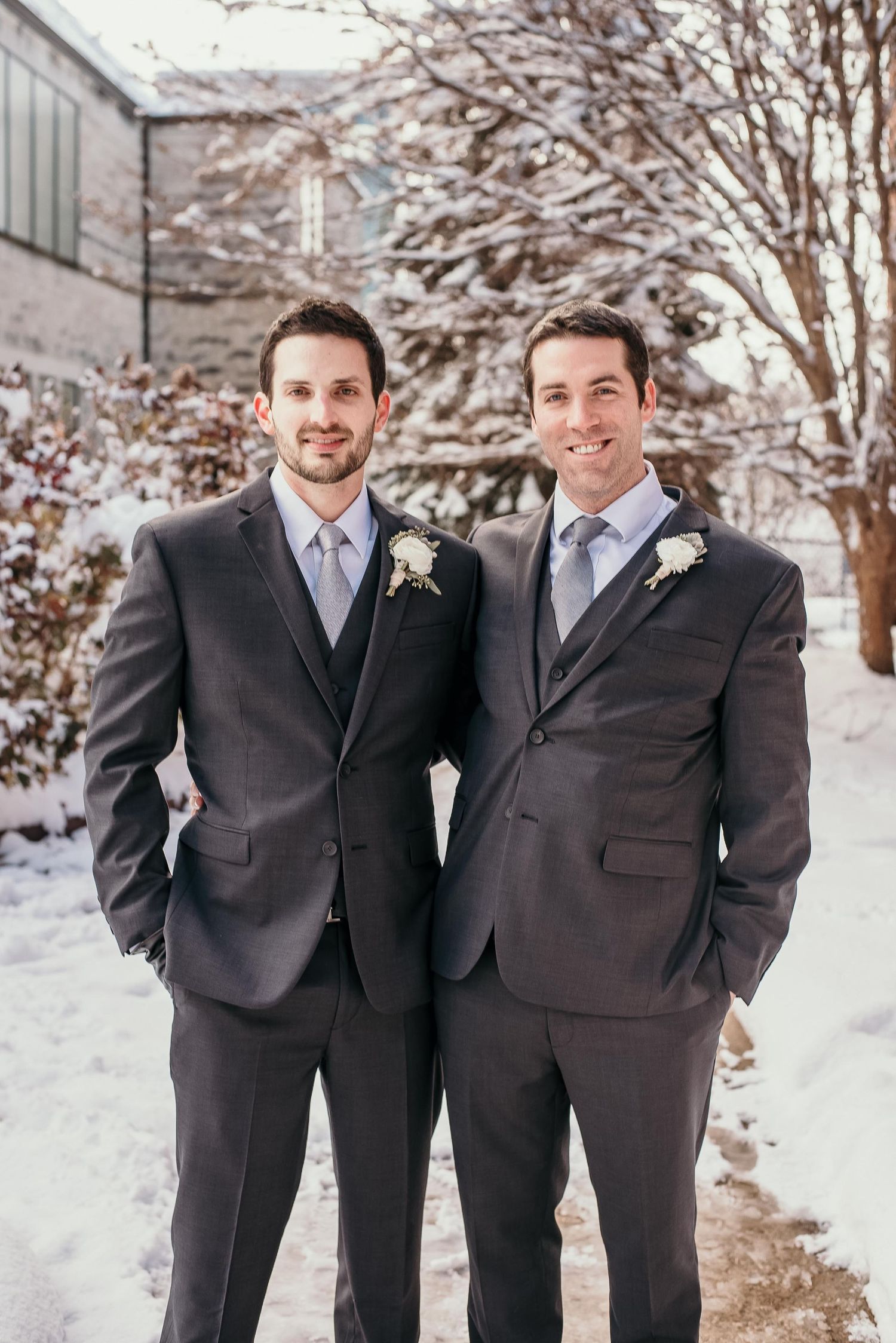 Two groomsmen in dark gray suits with boutonnieres posing outdoors in a snowy winter wedding setting.