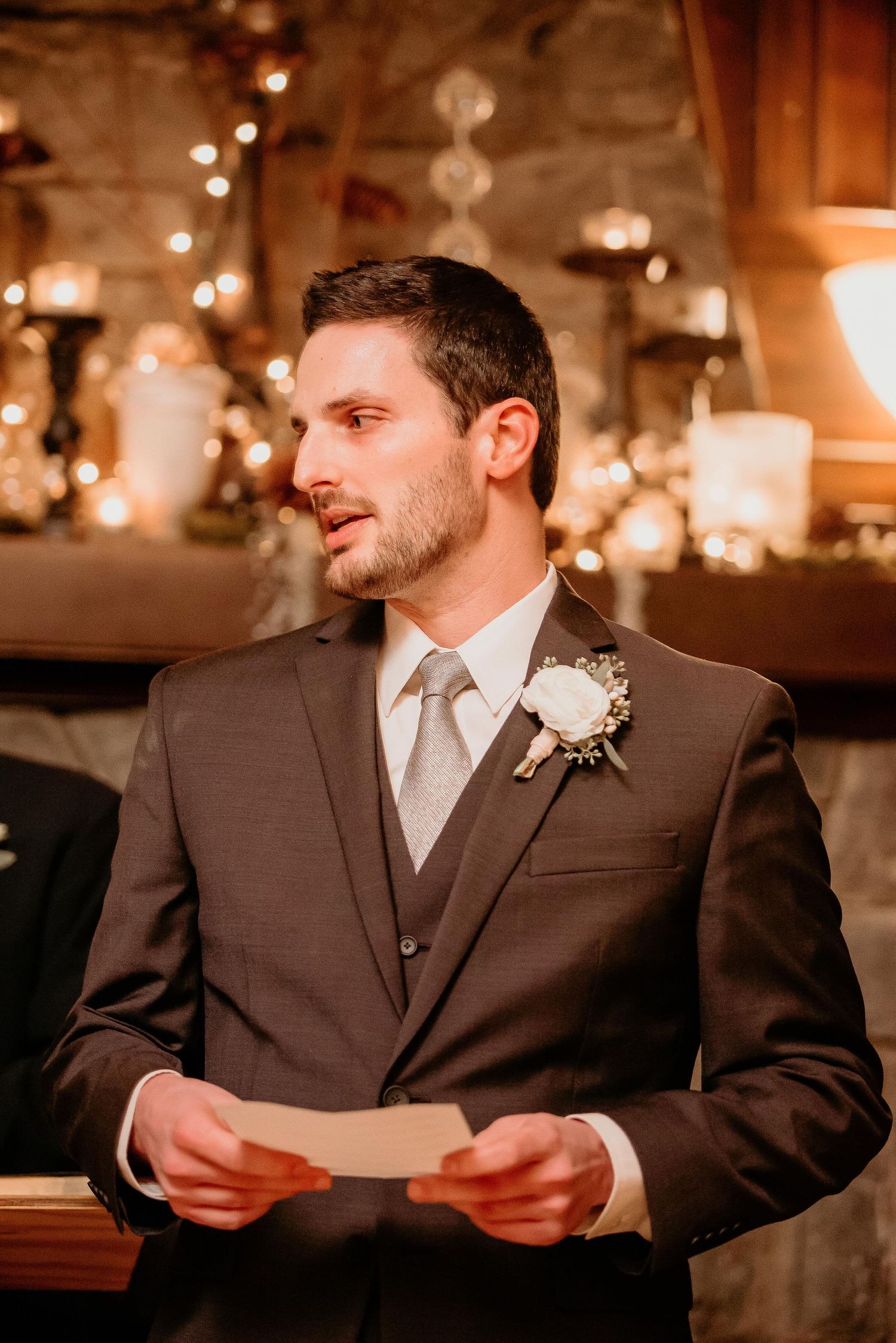 Groom in gray suit with white boutonniere reads vows at warmly lit wedding reception.
