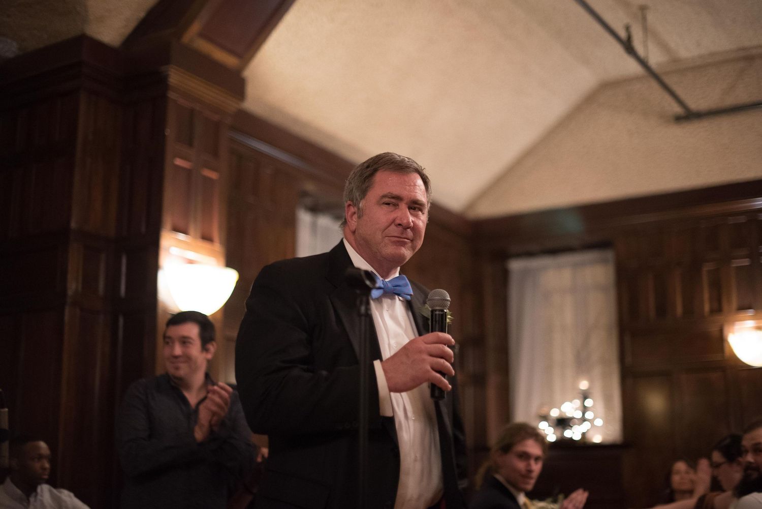 Man in tuxedo with blue bow tie speaks into microphone at formal evening event in elegant venue.