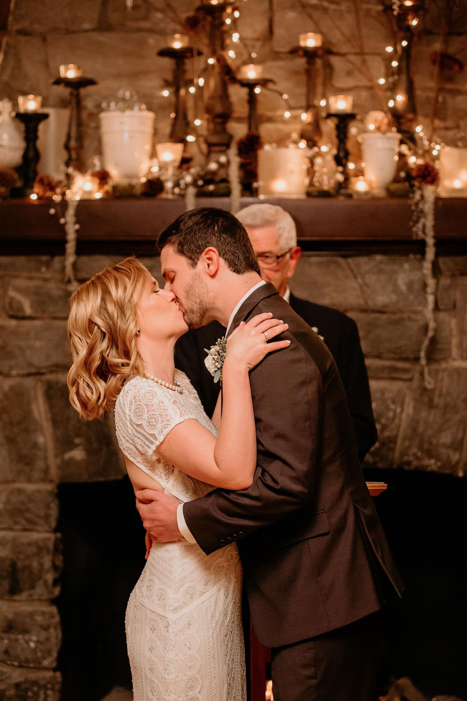 Bride and groom share first kiss at romantic stone fireplace wedding ceremony with candles and fairy lights.