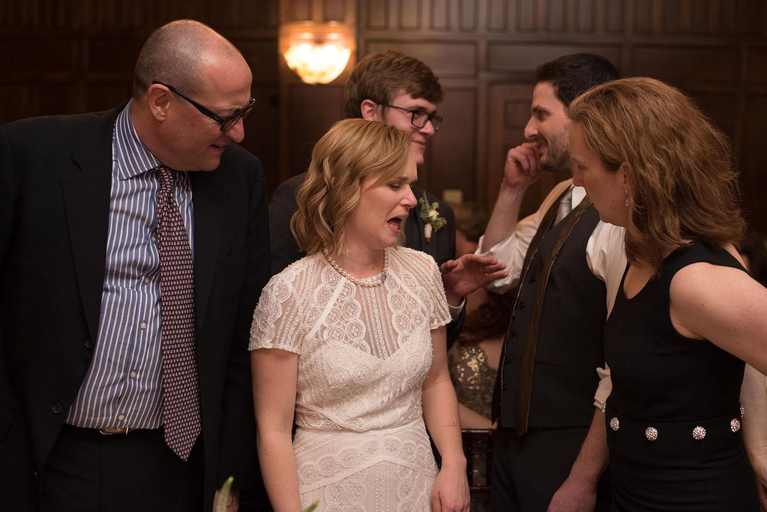 Guests in formal attire chat and laugh together at an elegant wedding reception in a wood-paneled venue.