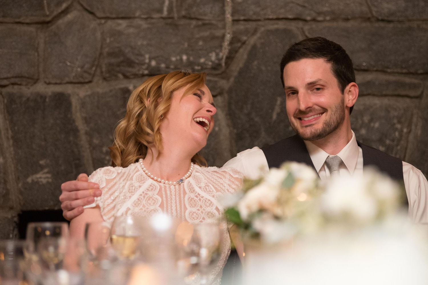 Bride and groom laughing joyfully at wedding reception table with stone wall backdrop and floral centerpiece.