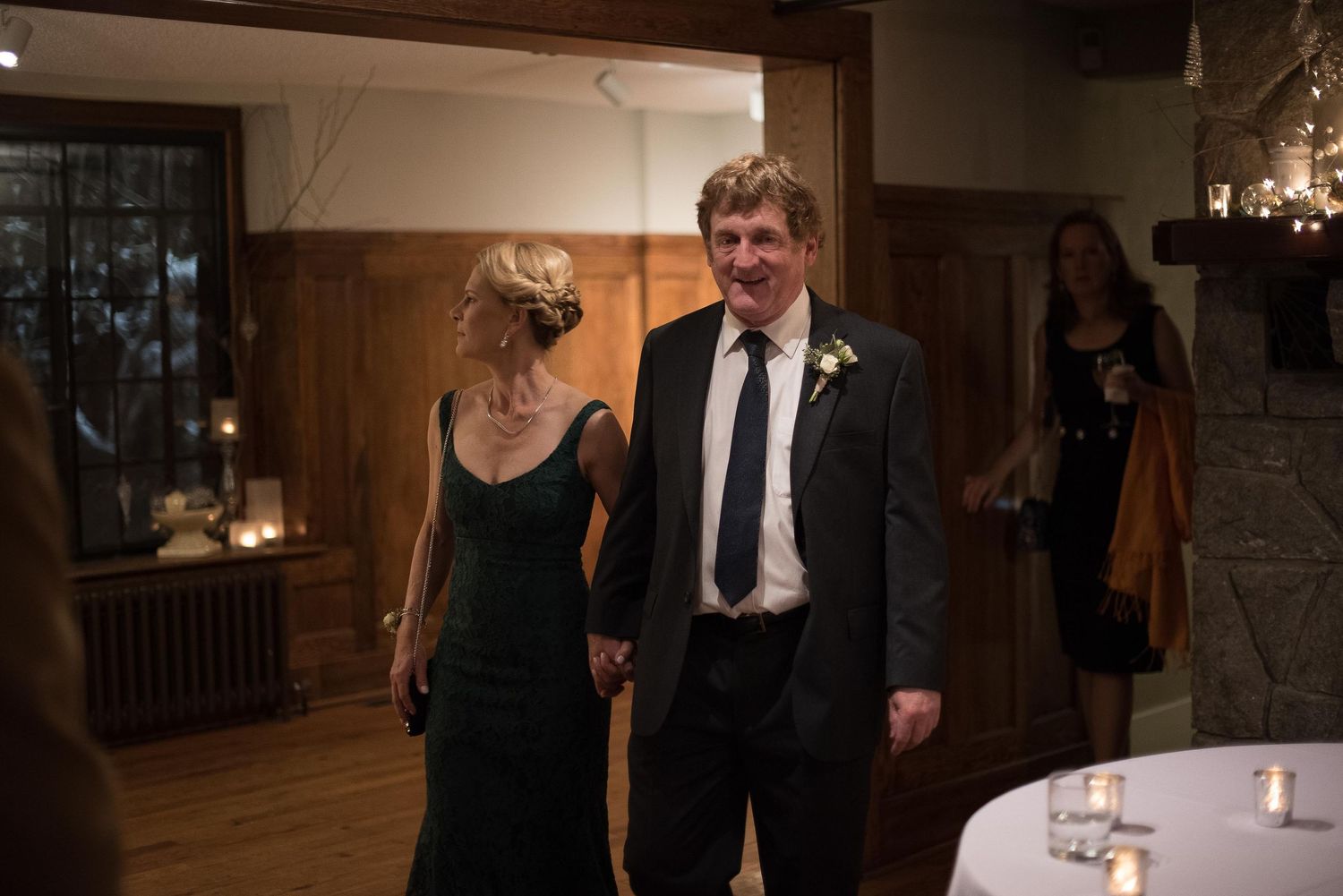 Elegant couple holding hands at a formal indoor event, man in suit with boutonniere, woman in black dress.