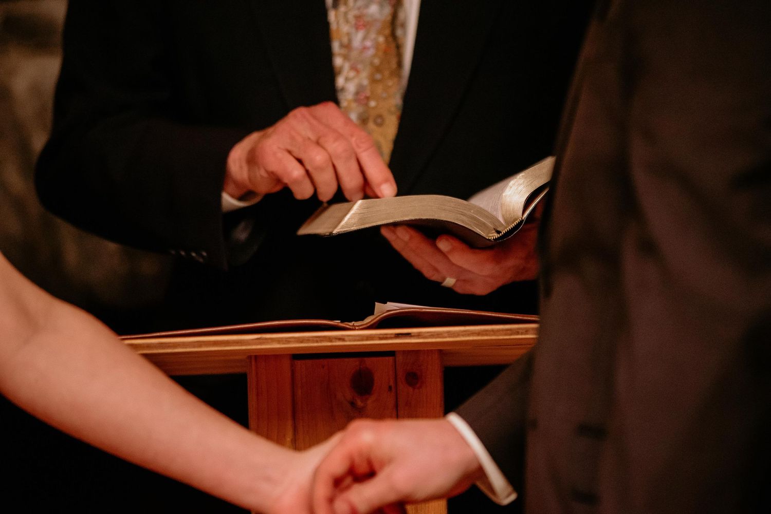 Officiant holding open Bible during wedding ceremony at wooden podium with couple's hands visible.