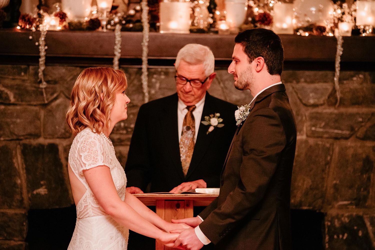 Bride and groom hold hands during intimate indoor wedding ceremony before stone fireplace with officiant.