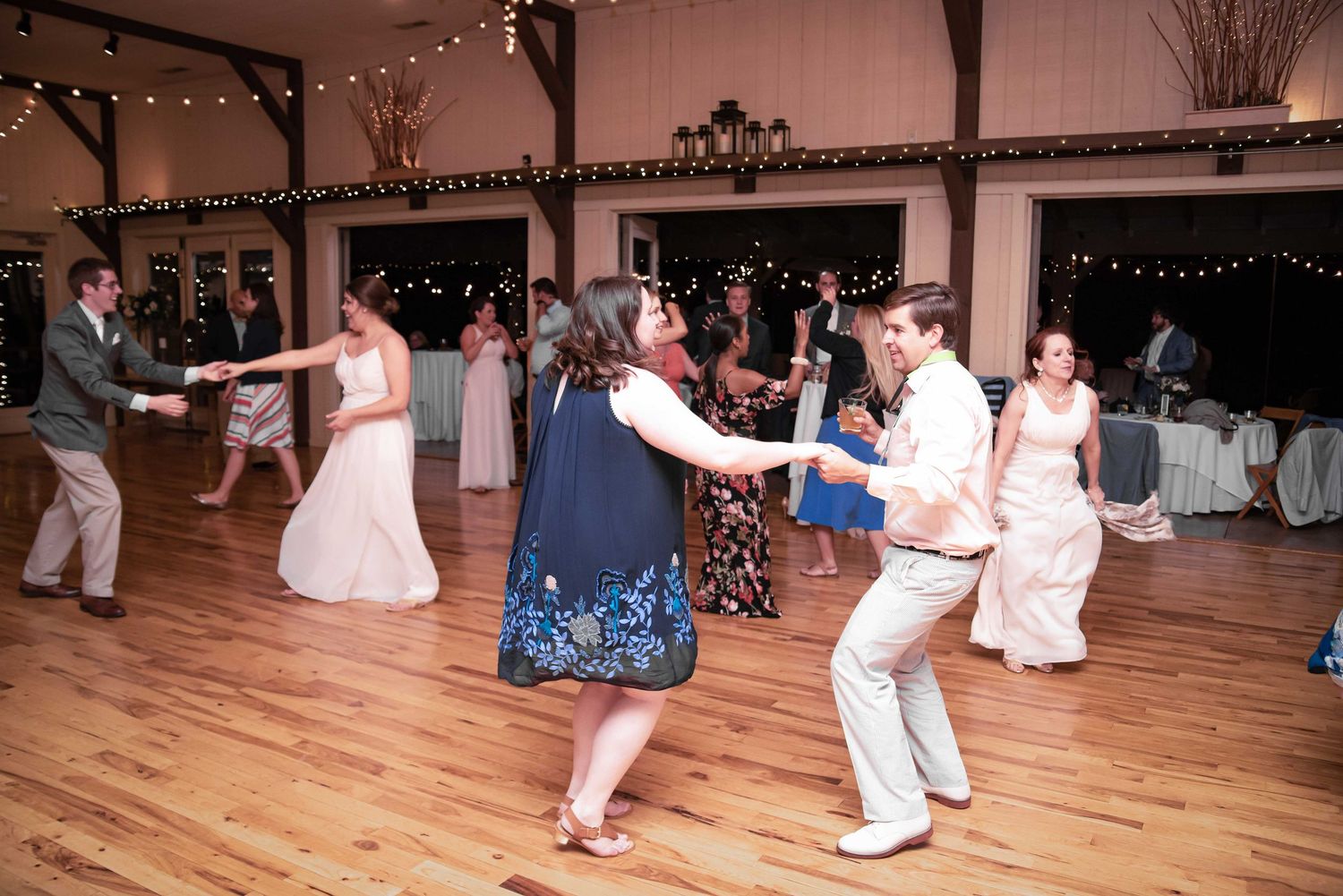 Guests dancing joyfully at a rustic wedding reception on a wooden dance floor with string lights.