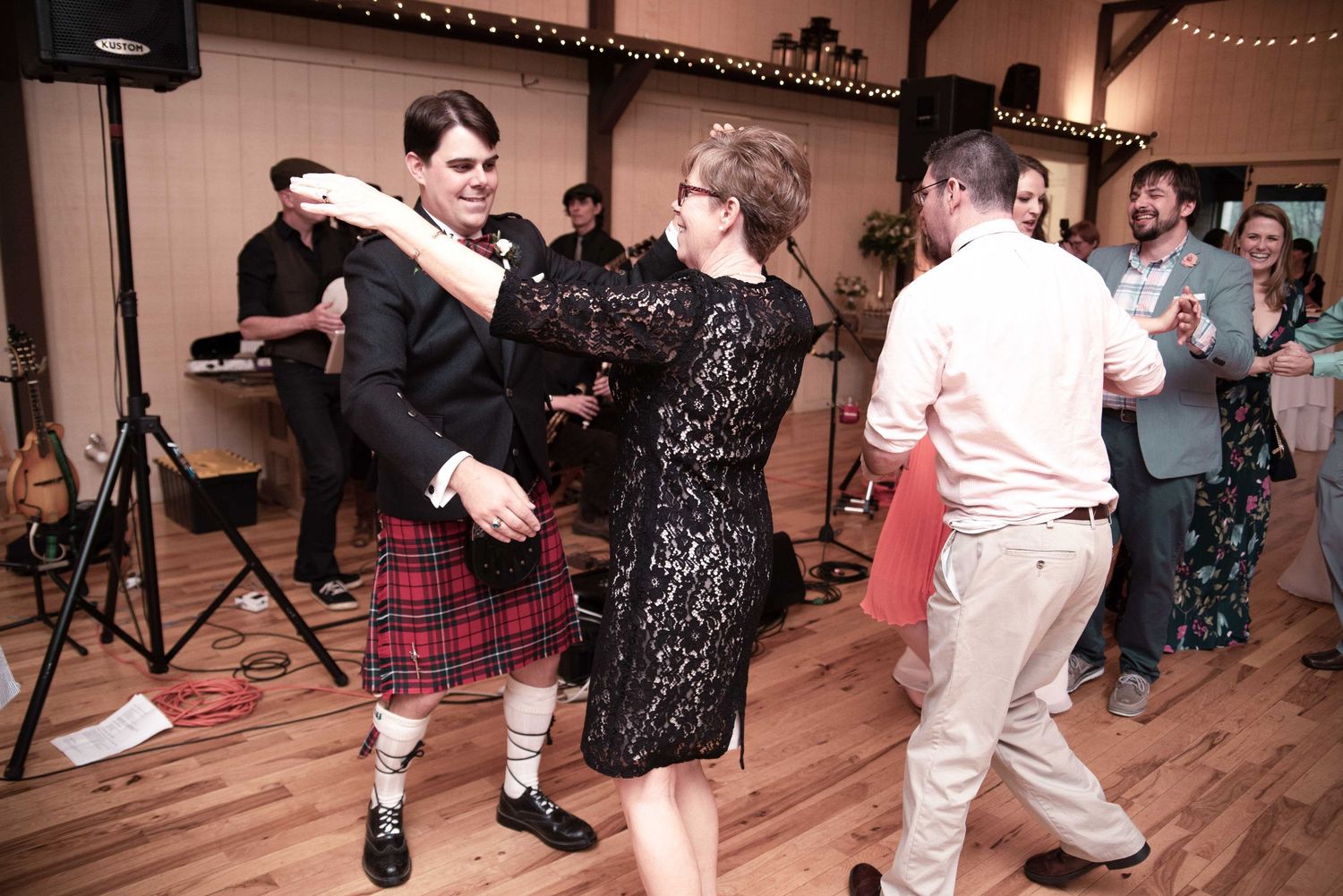 Guests dancing at a wedding reception, man in Scottish kilt and woman in black lace dress, live band performing in background.