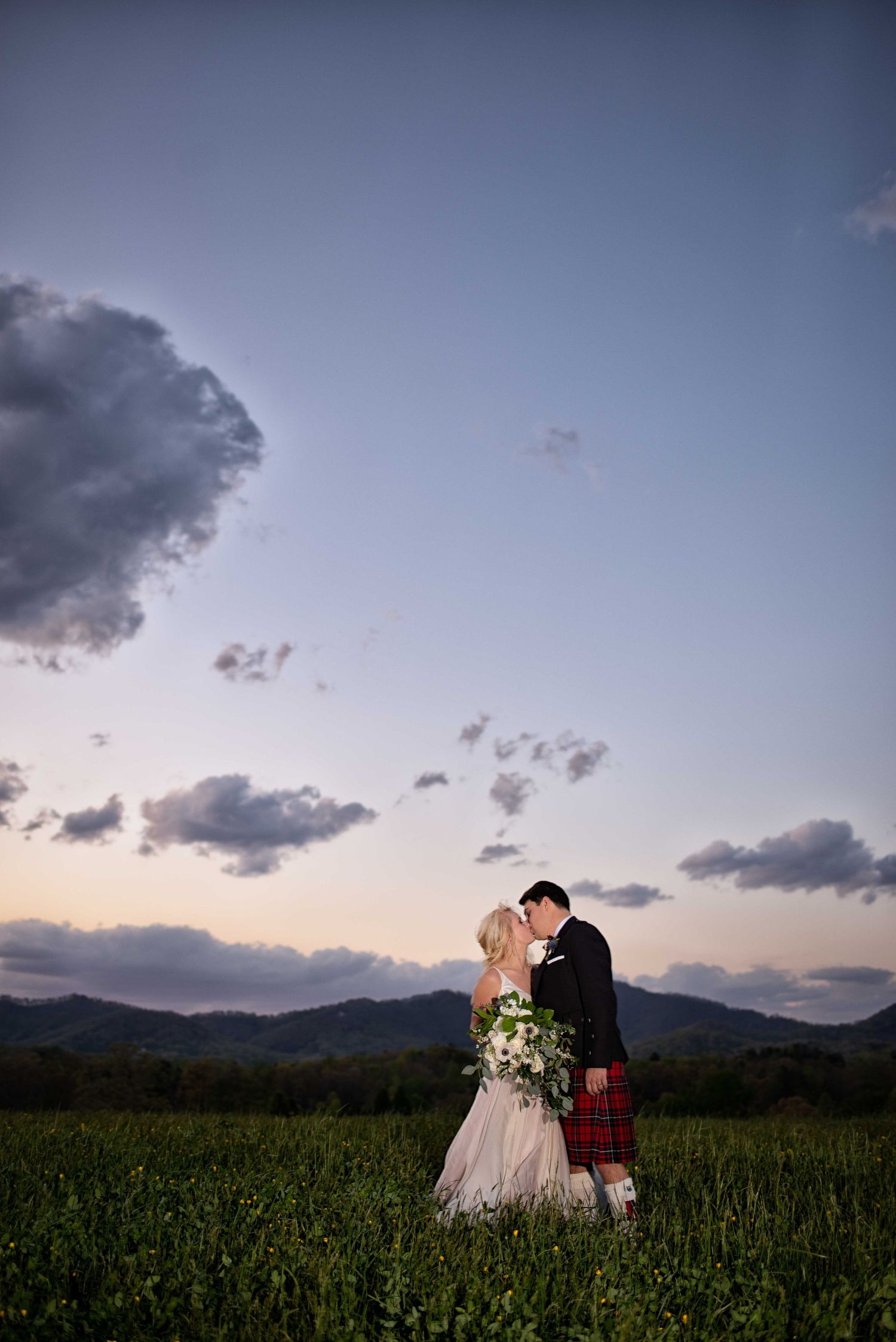 Bride and groom share a kiss at sunset in a green field with mountain backdrop, groom wearing red tartan kilt.