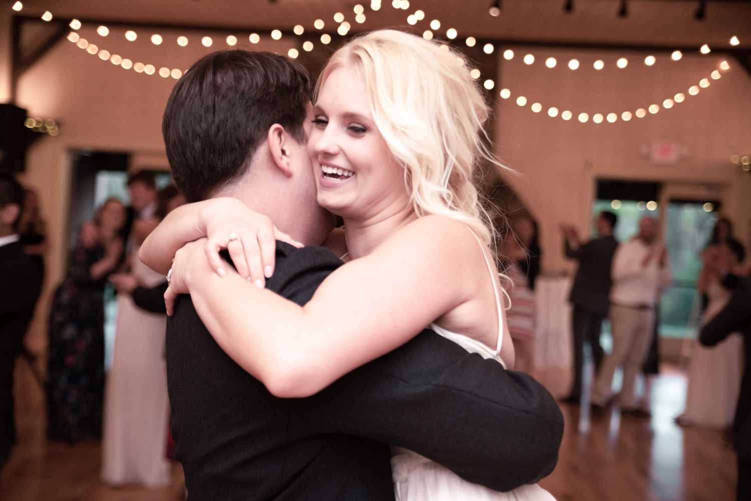 Couple sharing a romantic first dance embrace at a wedding reception with warm string lights glowing in the background.