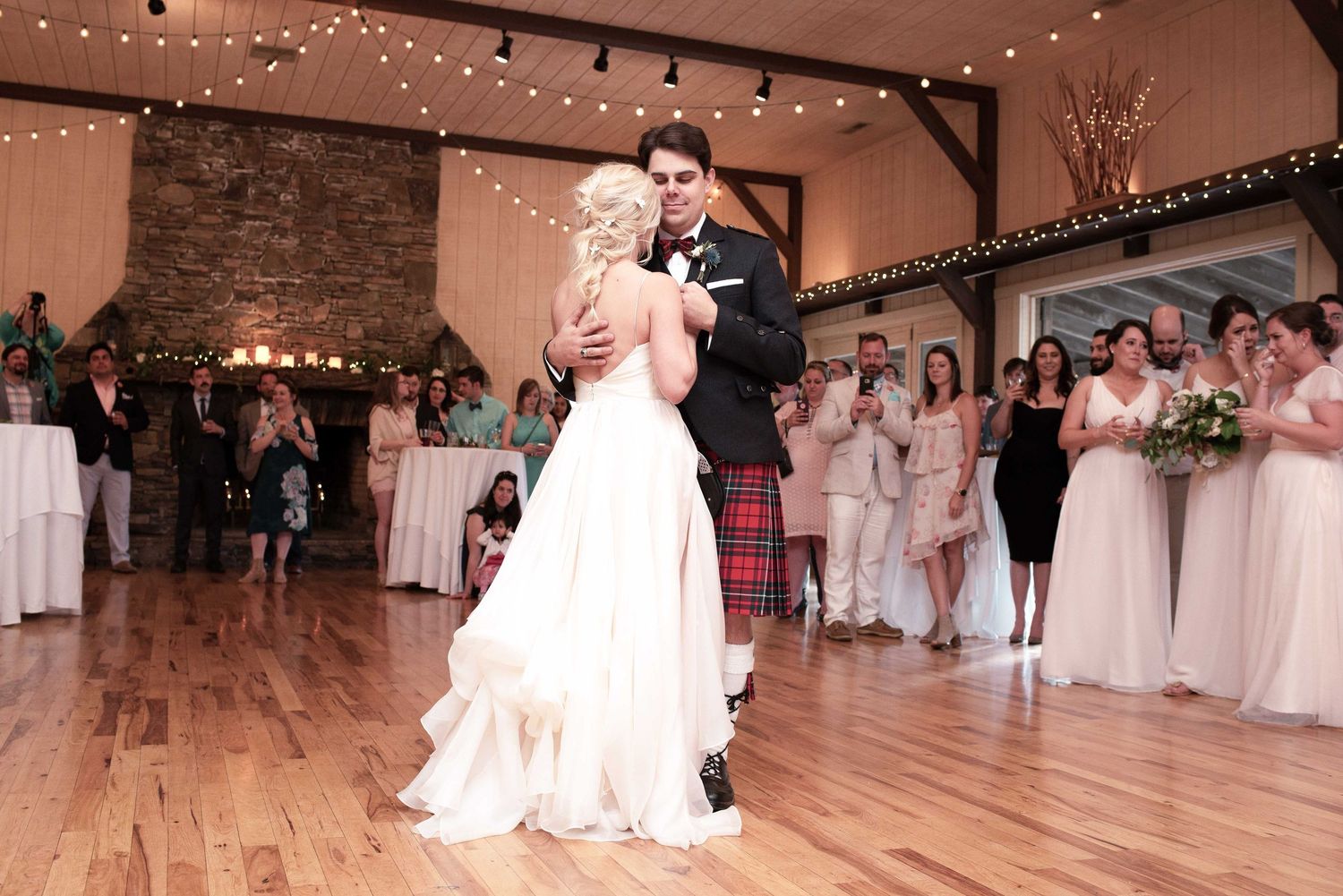 Bride and groom share first dance on wooden floor in rustic barn venue with string lights and wedding guests watching.