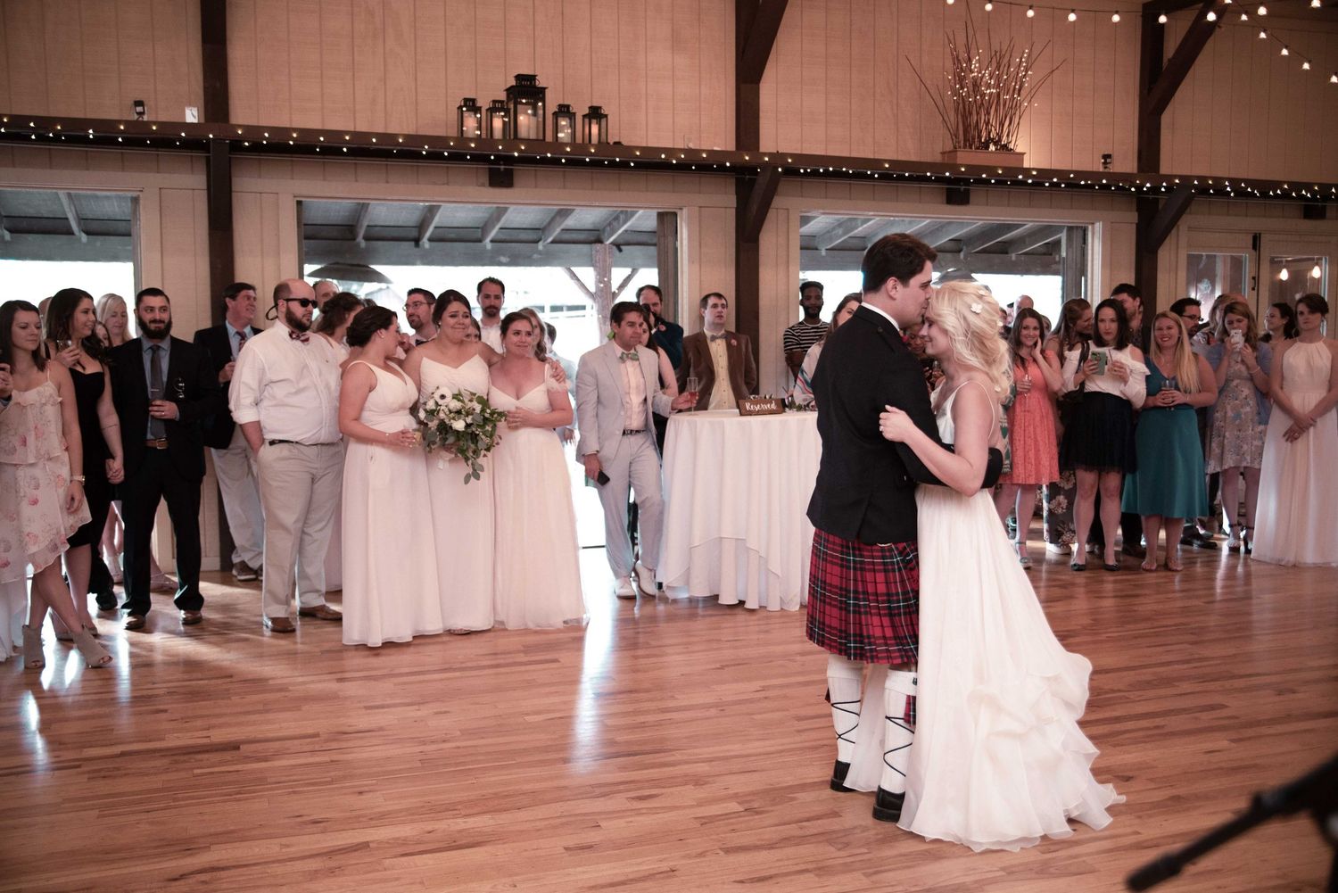 Bride and groom share first dance in rustic barn wedding venue with guests watching in background.