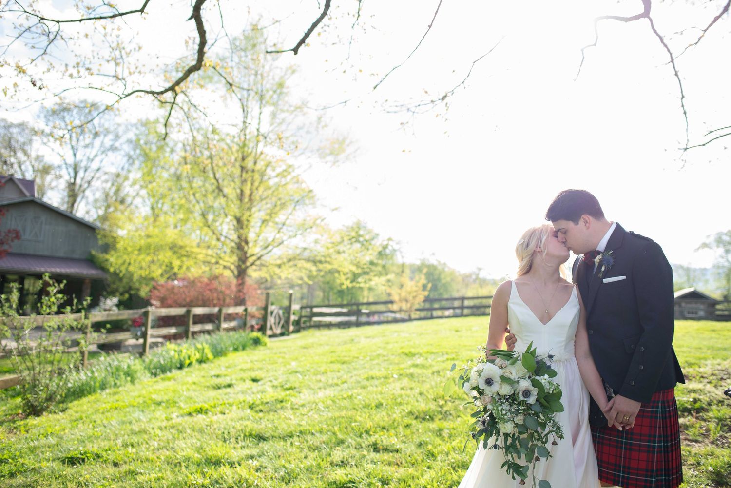 Bride and groom share a tender moment in a sunlit green field, she holds white bouquet, he wears a red tartan kilt.