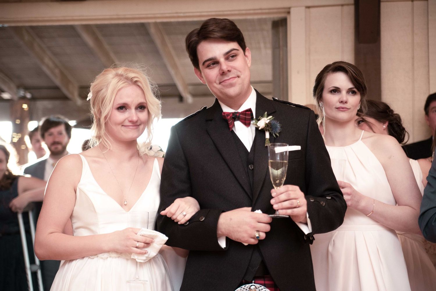 Bride and groom smiling at wedding reception, holding champagne glasses, with bridesmaid in white dress beside them.