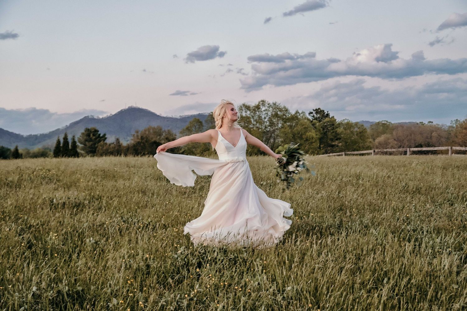 Bride in white wedding dress twirling in green meadow with mountain backdrop at sunset.