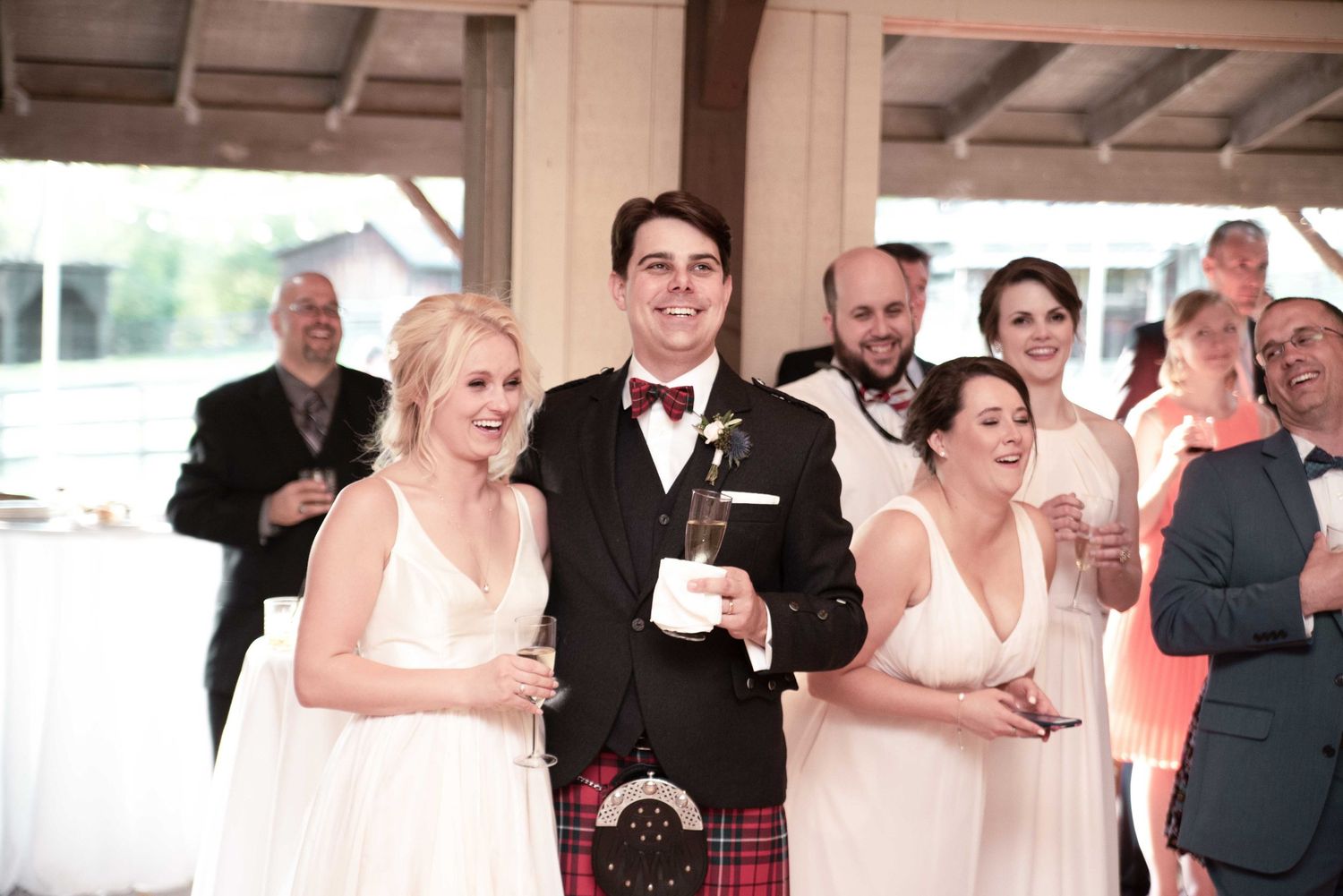 Groom in tartan kilt and bow tie smiles with bridesmaids in white dresses at a wedding reception.