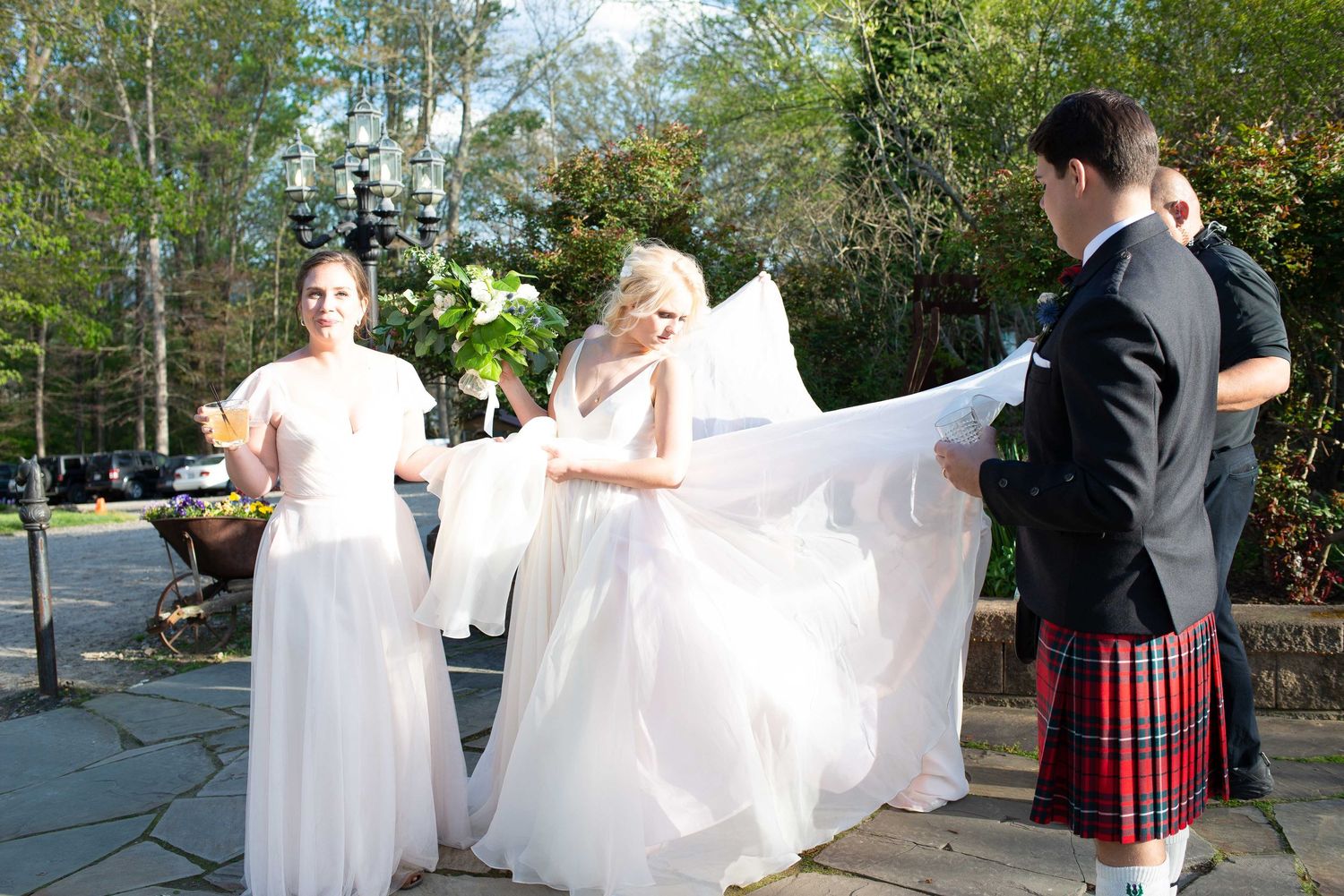 Bride in white gown with veil held by attendants, groom in red tartan kilt, outdoor wedding ceremony.