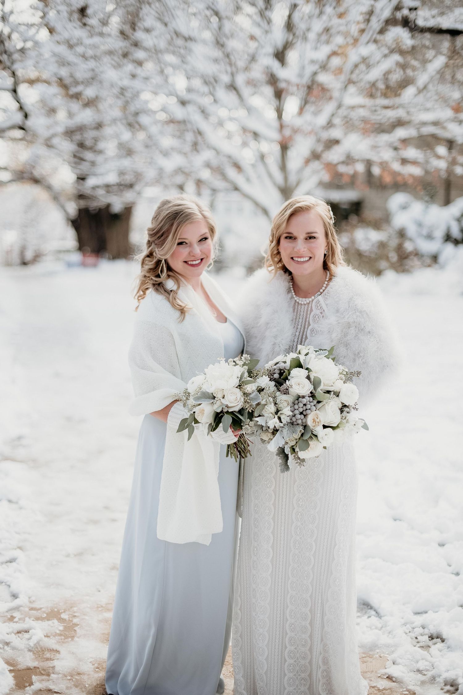 Two brides in white winter wedding attire holding floral bouquets, posing outdoors in a snowy landscape.