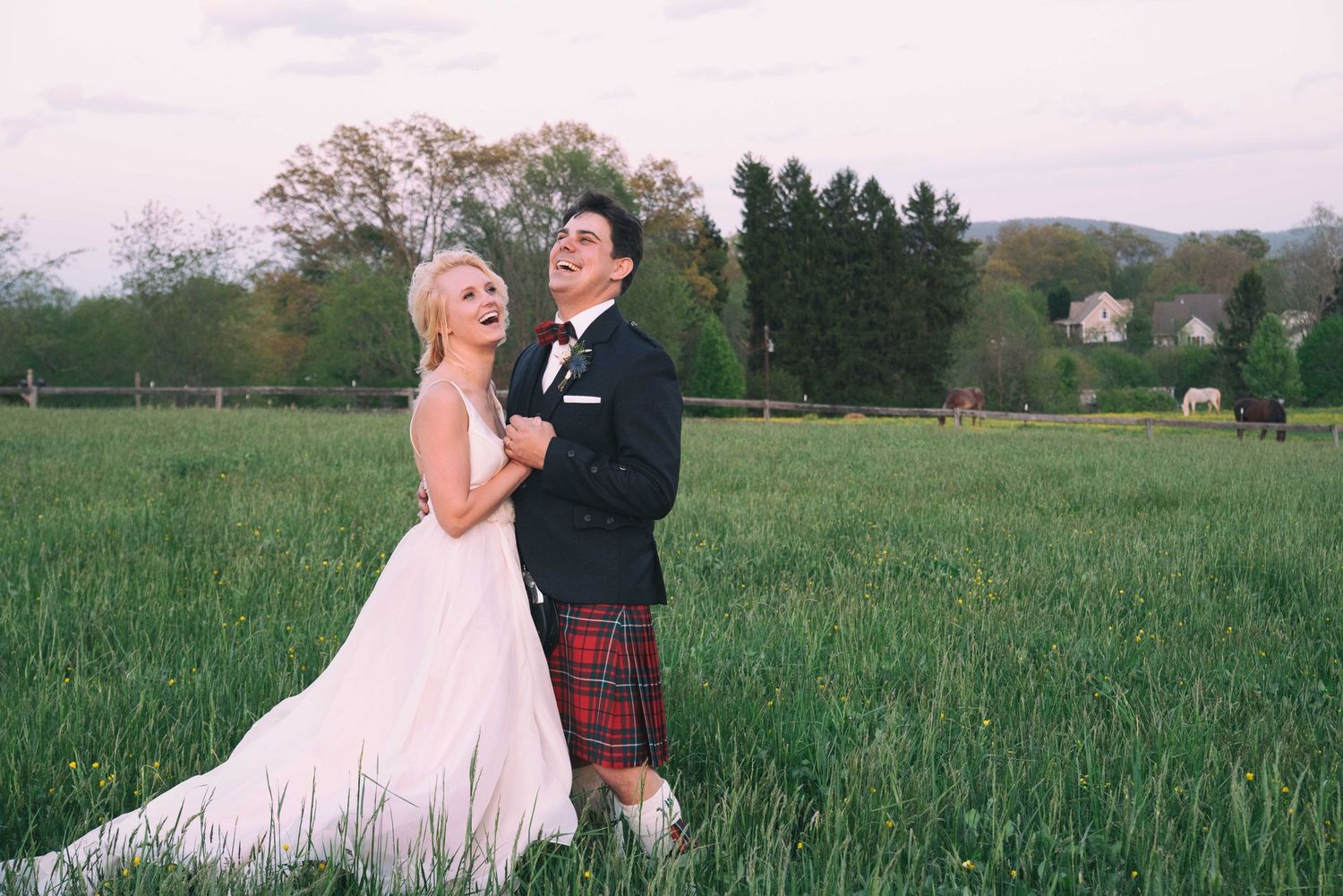 Bride in white gown and groom in tartan kilt laughing together in a lush green field at dusk.