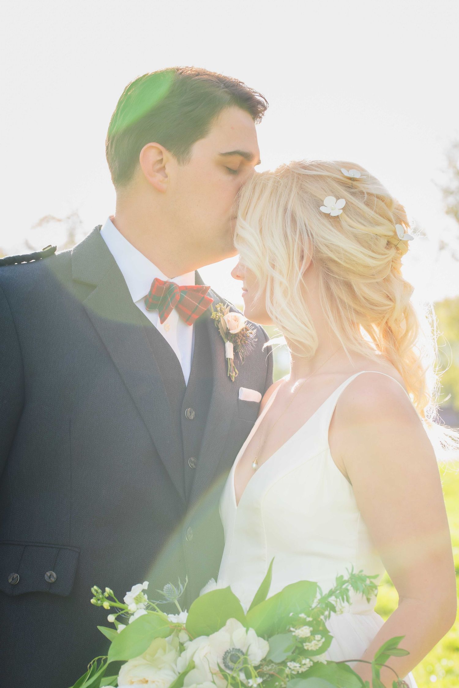 A bride and groom share a romantic kiss in golden sunlight, she holds a white floral bouquet.
