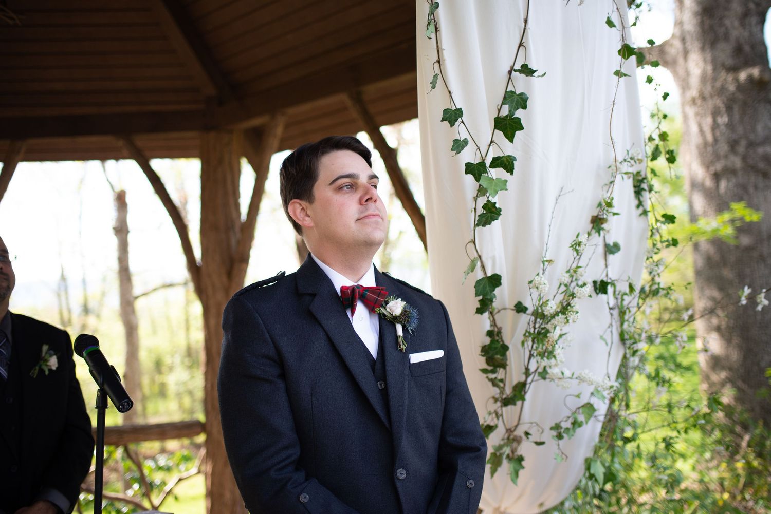 Emotional groom in navy suit and tartan kilt watching bride walk down aisle at outdoor wedding ceremony.