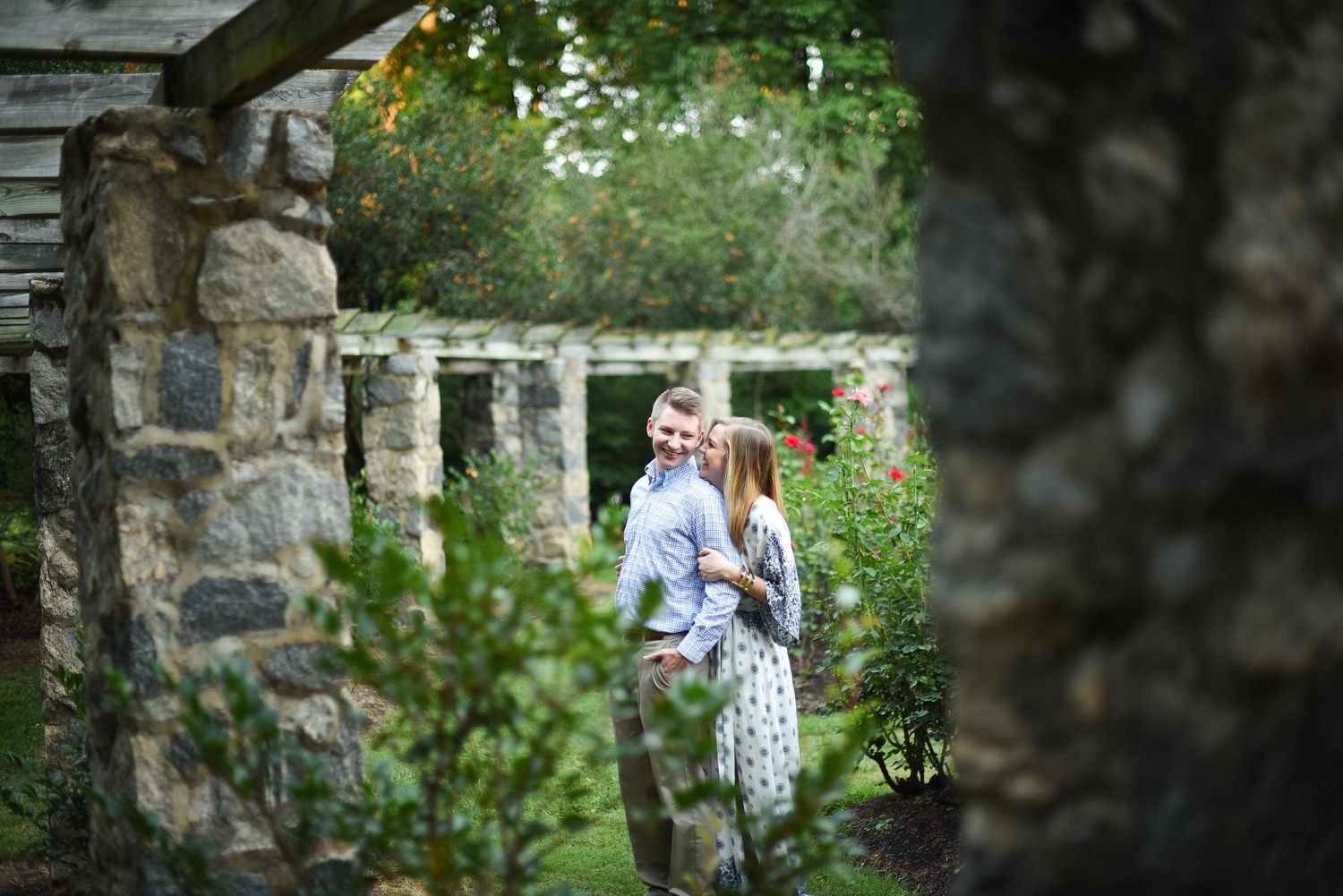 Couple embracing in a lush garden framed by stone archway and wooden pergola, surrounded by greenery.