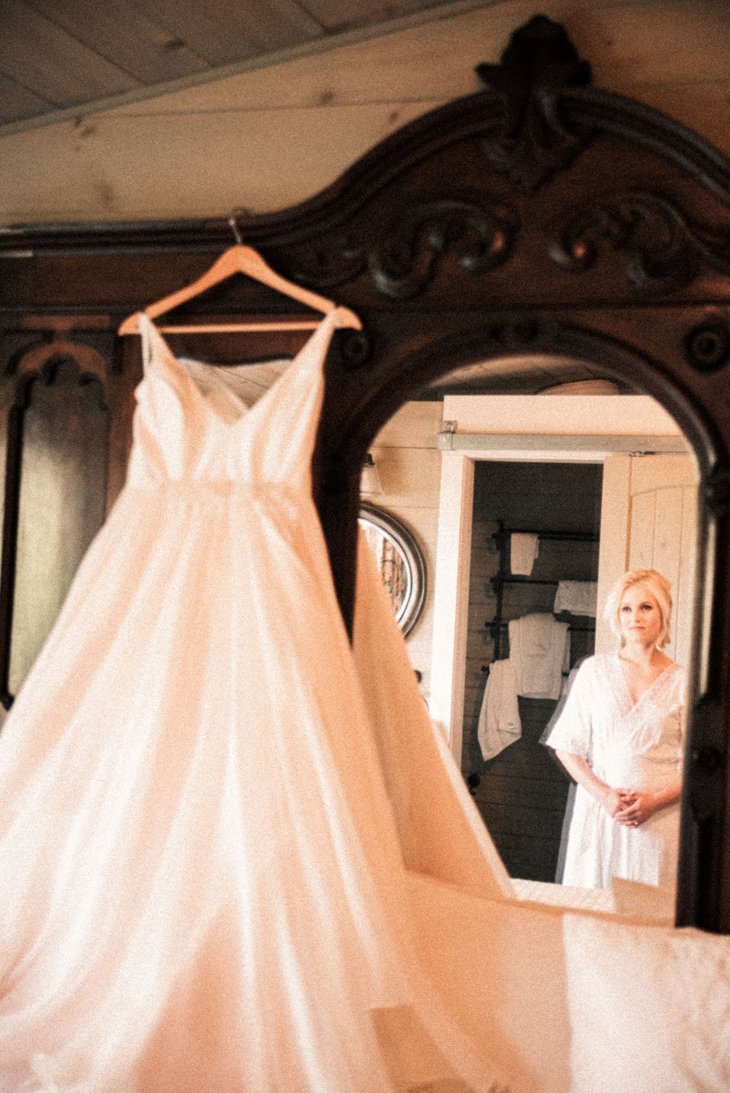 An elegant A-line wedding gown hangs on a mirror while the blonde bride admires it in the reflection.