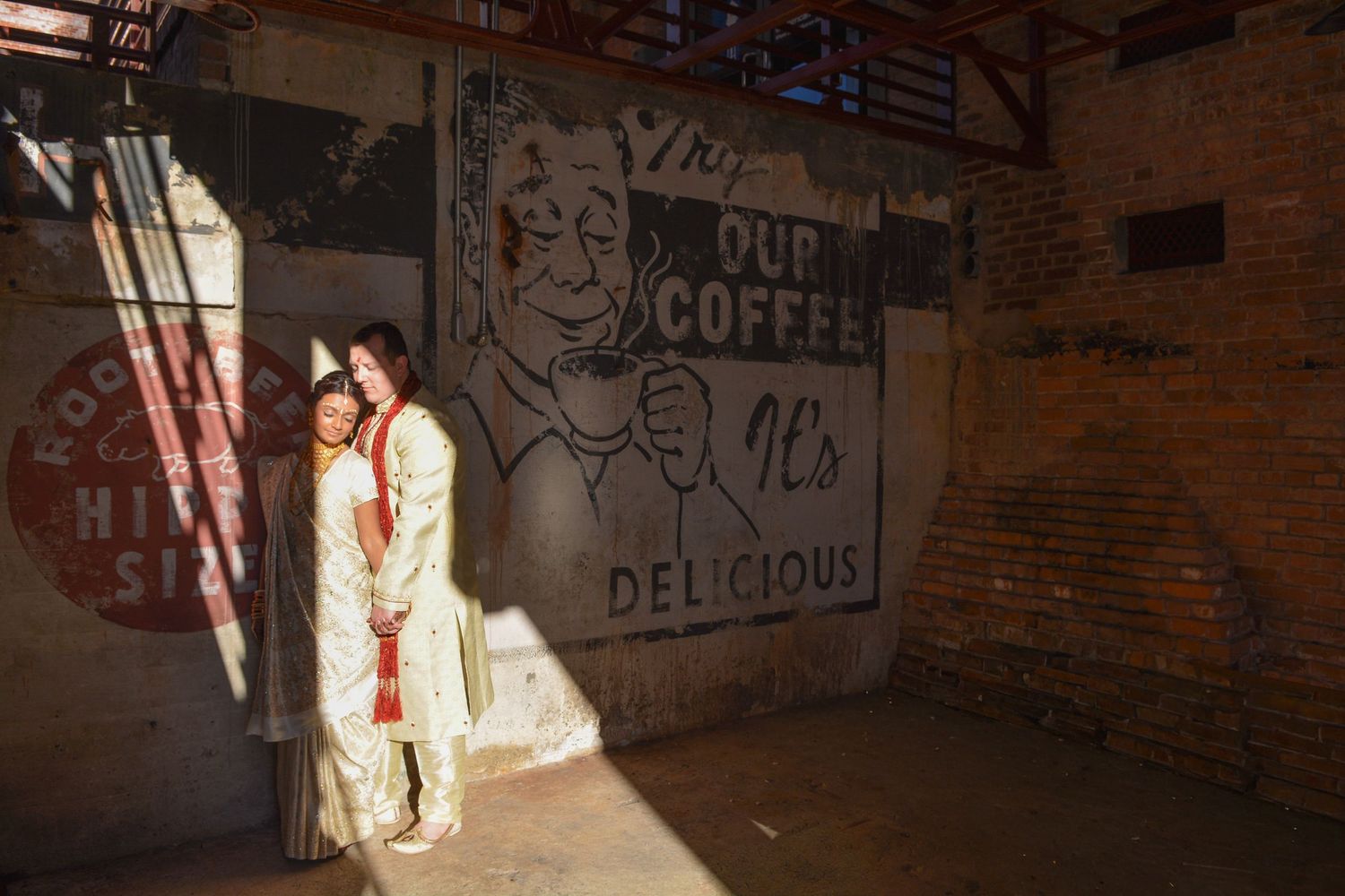 Couple in wedding attire posing before a vintage Our Coffee Its Delicious mural in a rustic brick building.