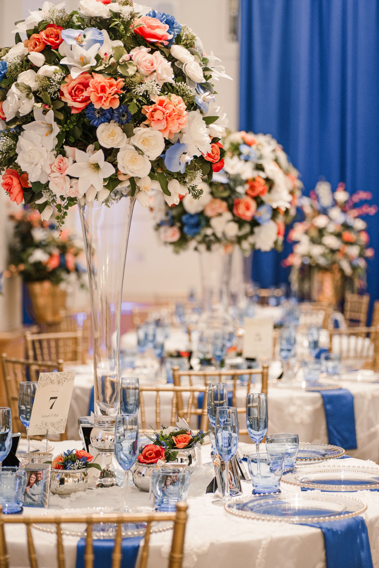 Elegant wedding reception table with tall floral centerpiece featuring coral and white blooms, blue napkins, and gold chairs.