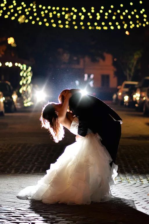 Bride and groom share a romantic dip kiss on cobblestone street at night, surrounded by glowing bokeh lights.