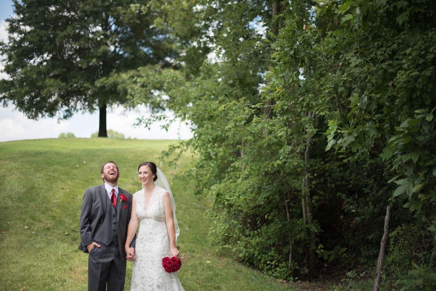 Bride and groom holding hands laughing outdoors on lush green hillside with trees on their wedding day.
