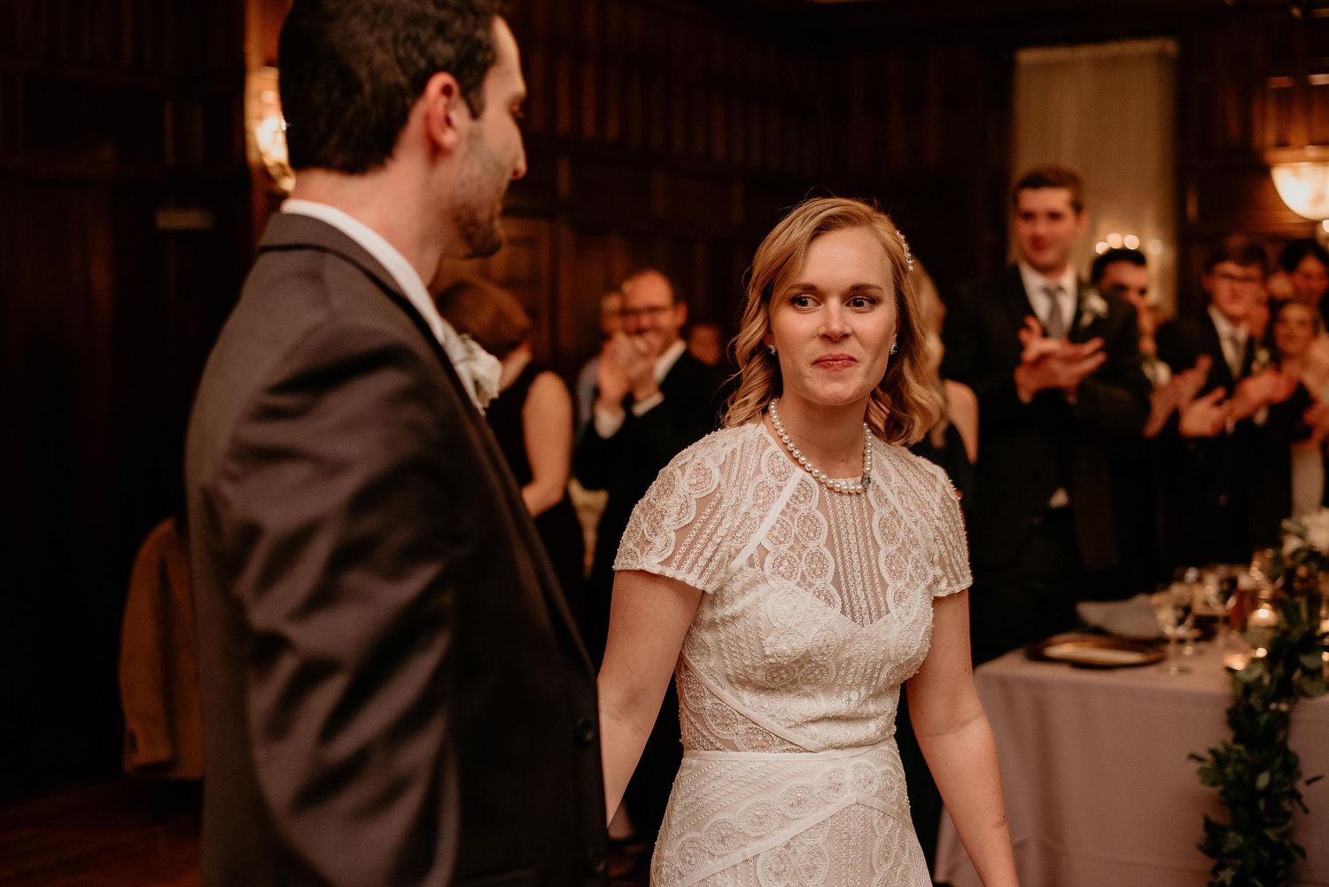 Bride in white lace dress holds groom's hand at wedding reception while guests applaud in elegant wood-paneled venue.
