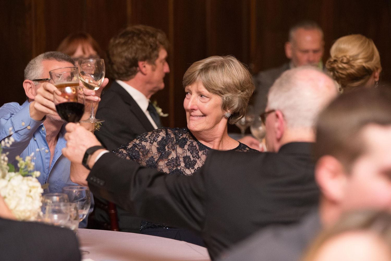Guests raising wine glasses in a toast at an elegant formal dinner event, with a smiling woman in black lace dress.