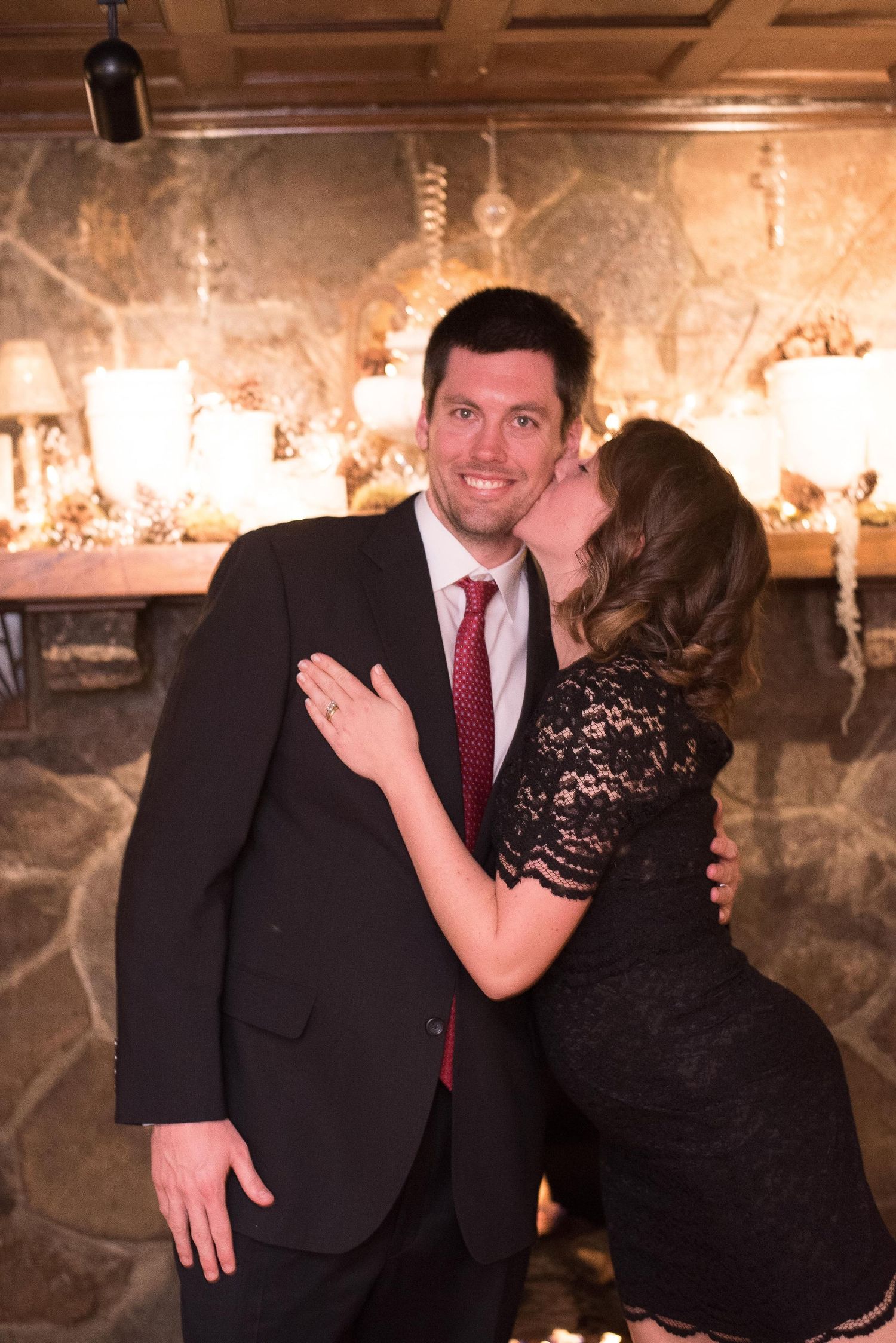 Couple posing by stone fireplace, woman in black lace dress kissing man in dark suit with red tie.