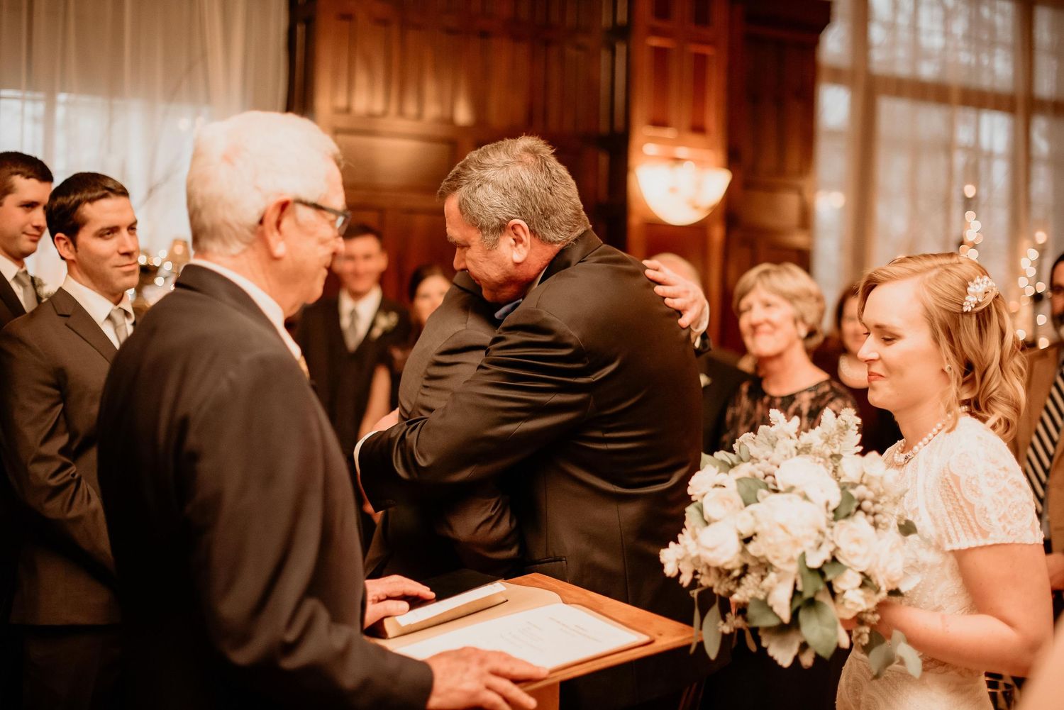 Groom embracing a guest at wedding ceremony while bride holds white floral bouquet indoors.