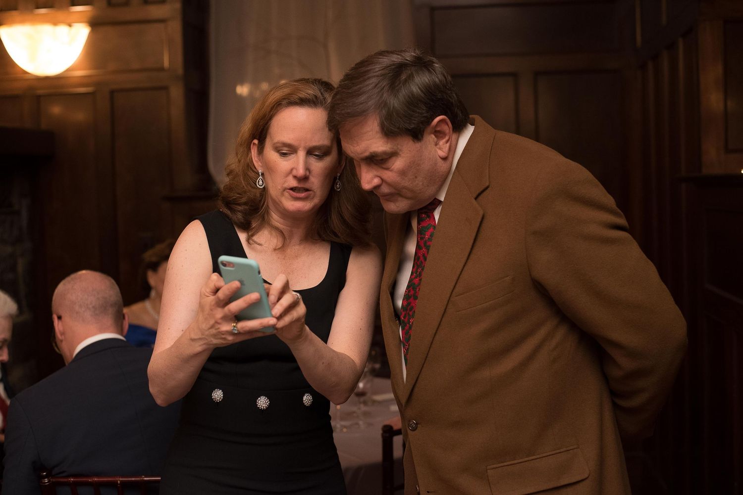 A woman in a black dress shows her smartphone to a man in a brown suit at a formal indoor event.