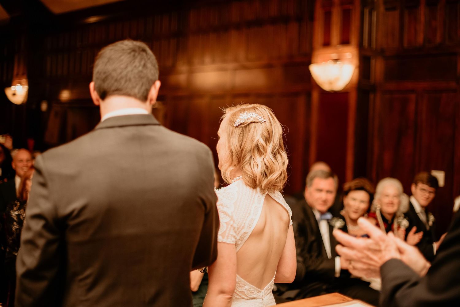 Bride and groom stand at wedding altar facing guests who applaud in elegant wood-paneled reception hall.