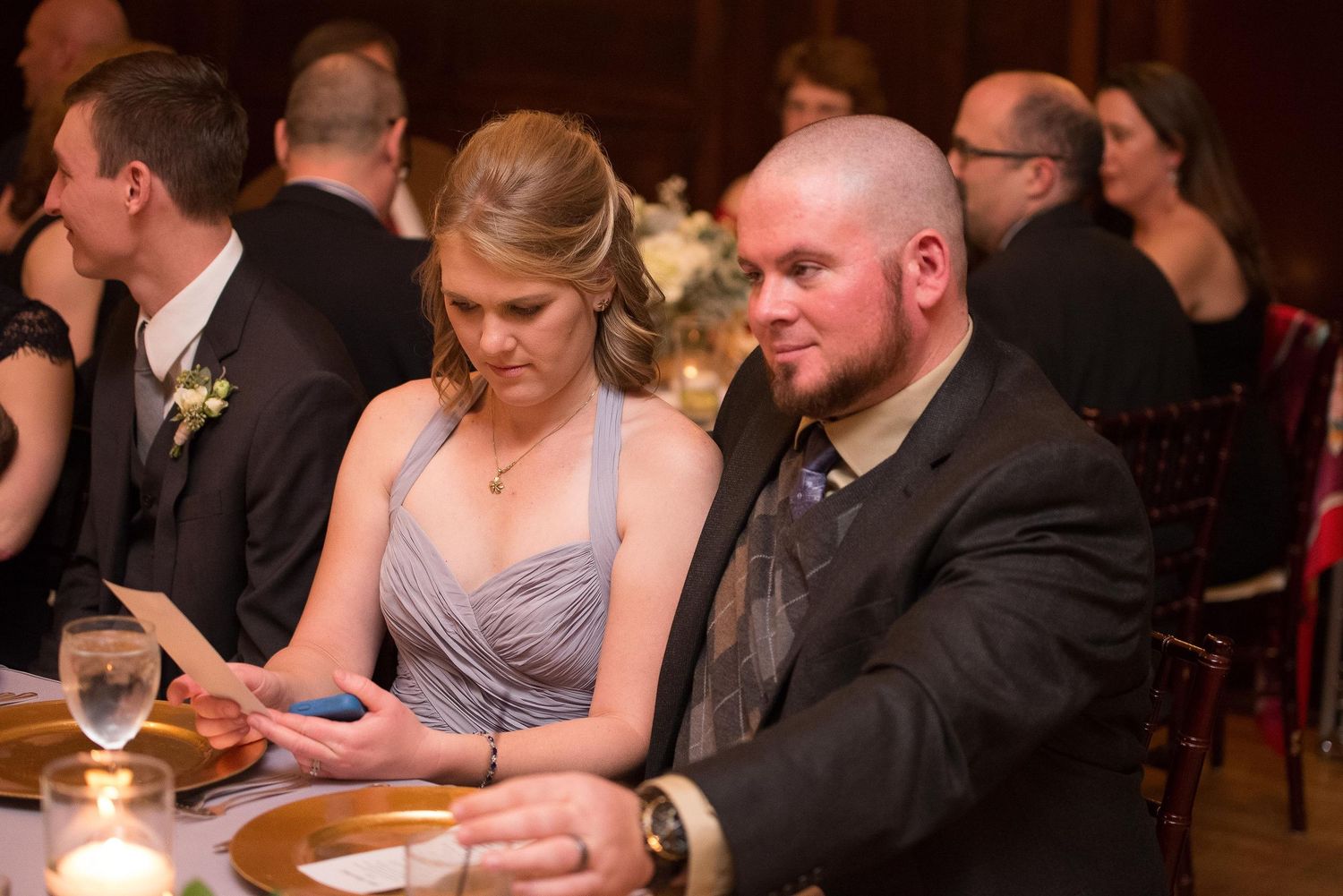 A man and woman in formal attire review a card together at an elegant wedding reception dinner.