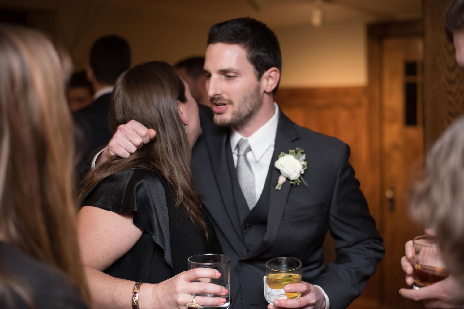Groom in black suit with white boutonniere holds drink while socializing with guests at wedding reception.