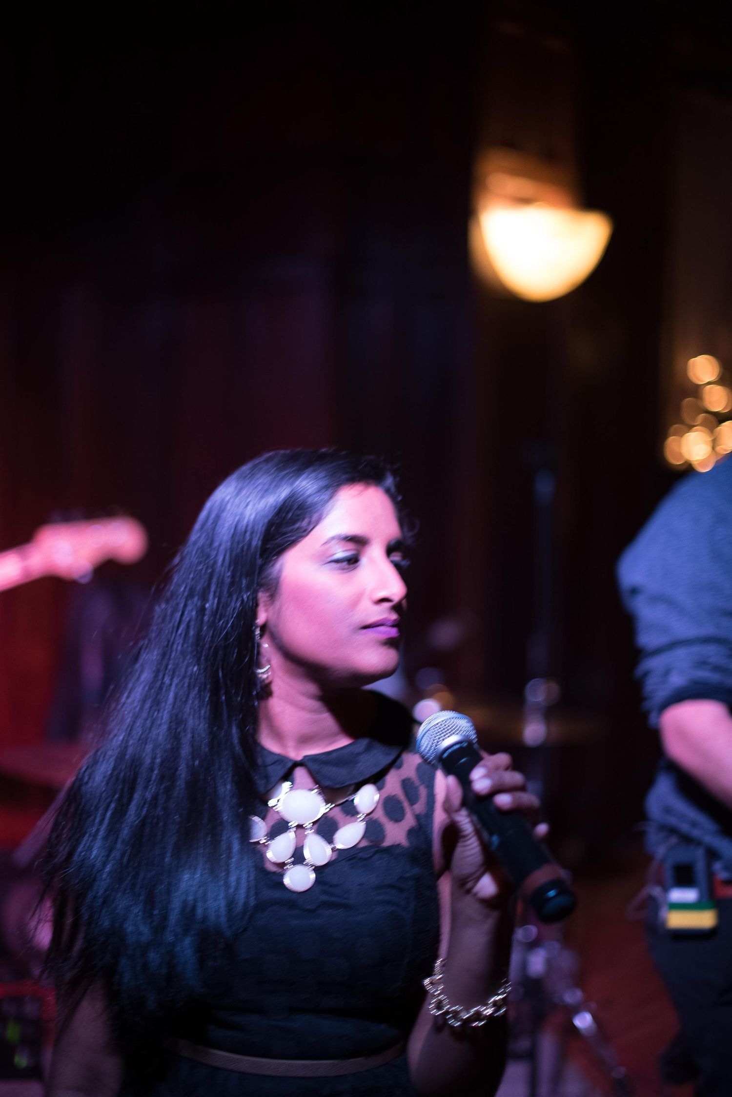 A woman with long black hair sings into a microphone on stage, wearing a black top and statement necklace.