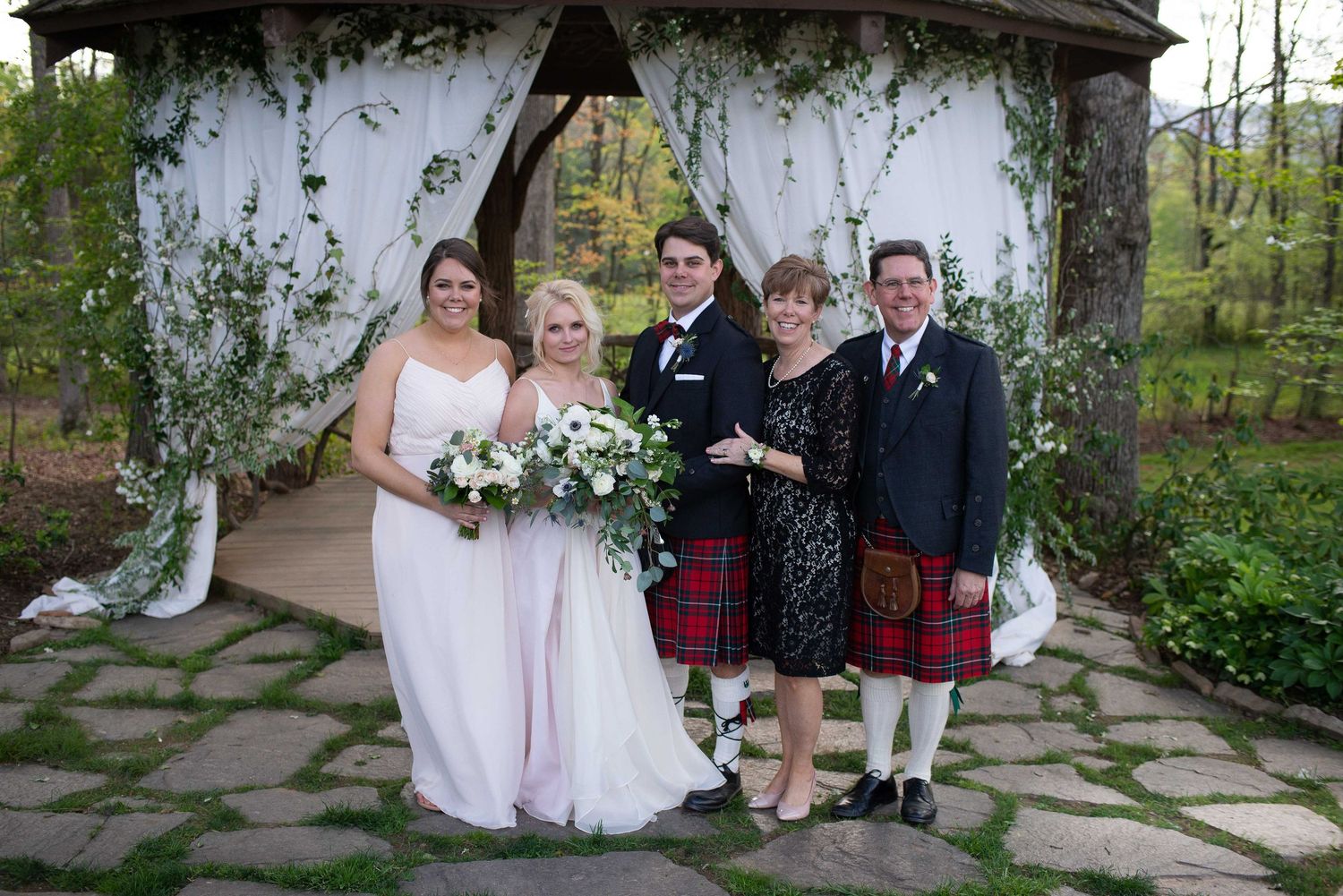 Wedding party poses under floral gazebo, bride in white, bridesmaid in pink, groom in tartan kilt, couple in formal wear.