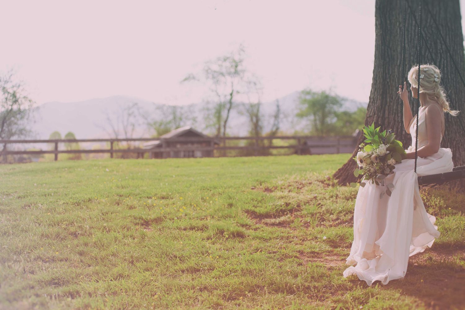Bride in white gown holding bouquet stands by large tree in rustic outdoor field with wooden fence and mountains.