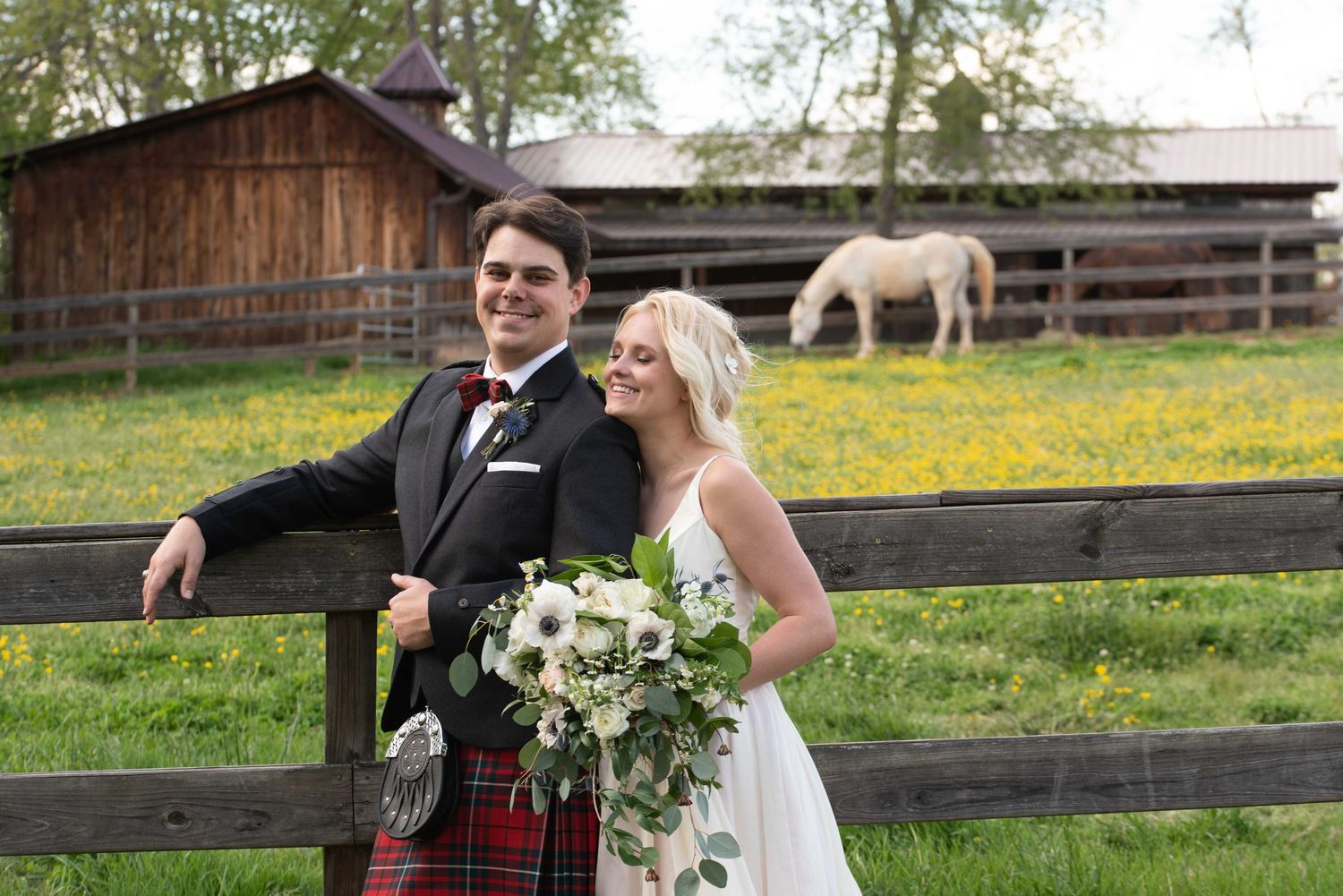 Bride and groom smiling by a wooden fence at a farm wedding, with a white horse grazing in a green field behind them.