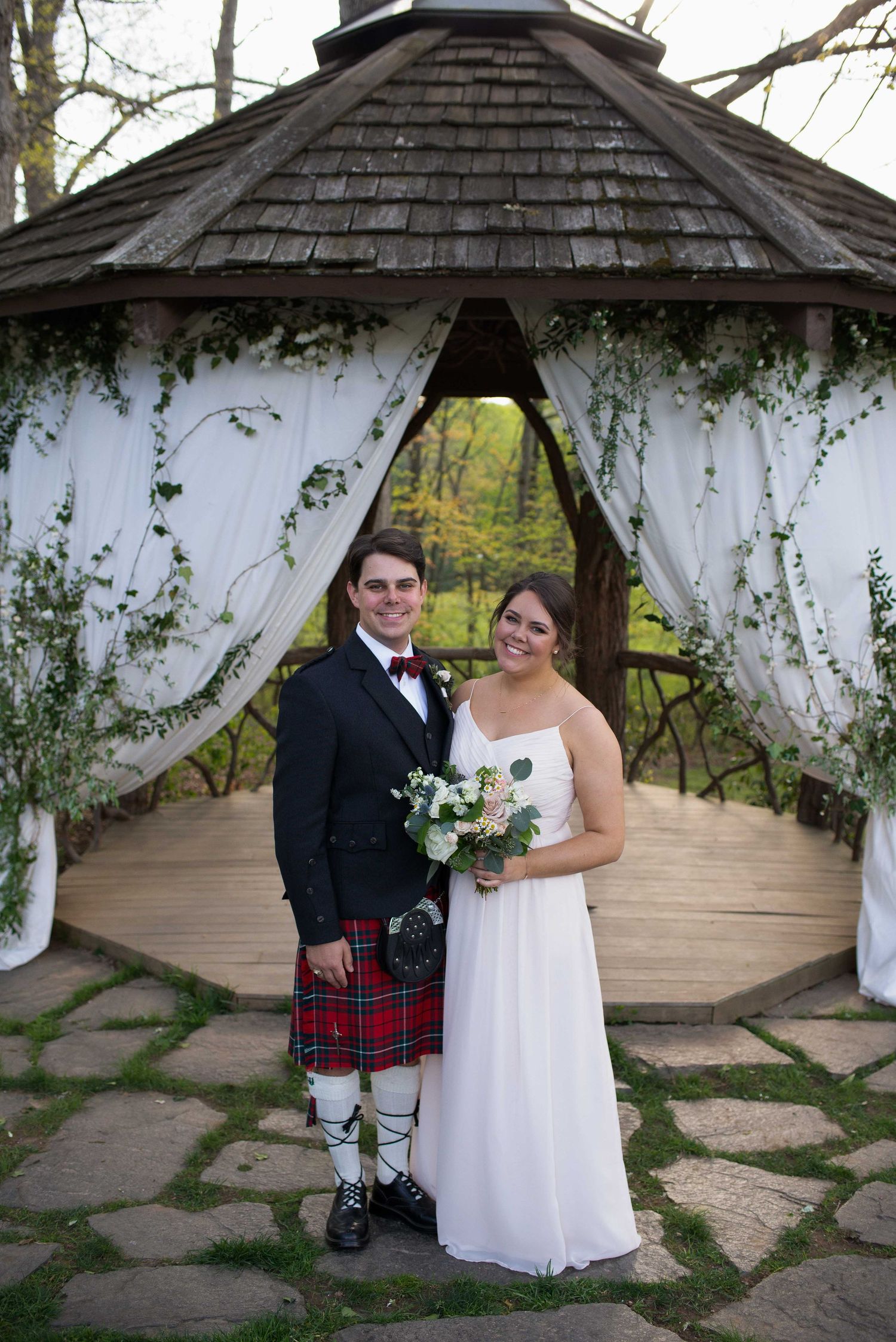 Bride and groom pose under decorated gazebo, groom wearing red tartan kilt and black jacket, bride in white gown holding bouquet.