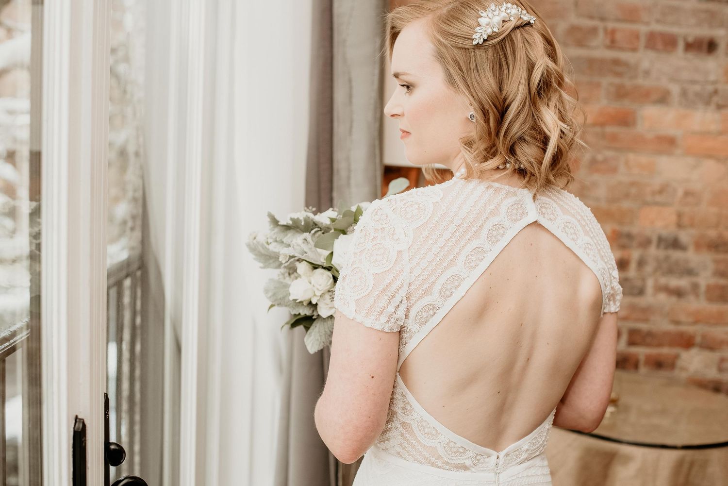 Bride in backless lace wedding gown holding white bouquet, standing by window with exposed brick wall.