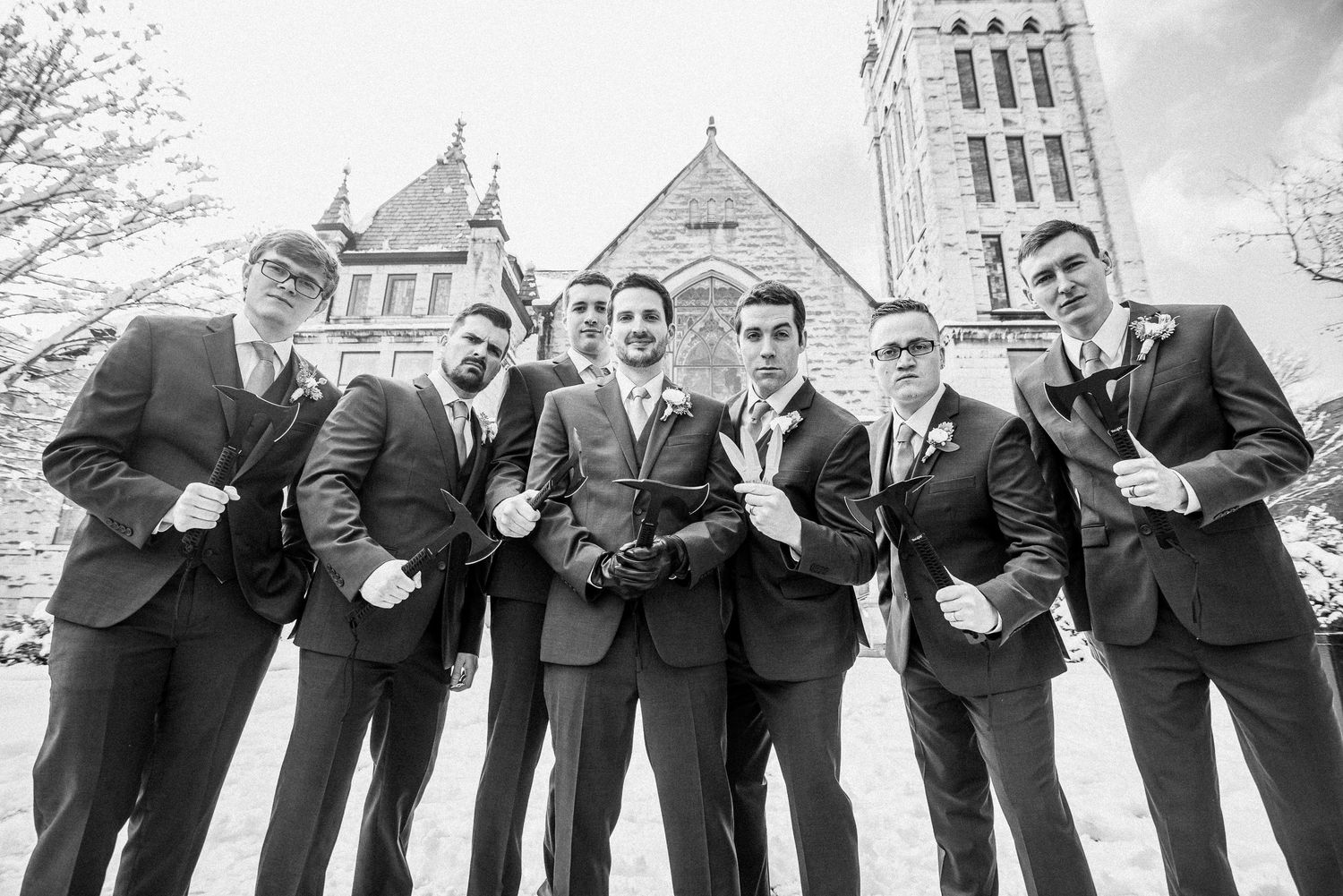 Black and white photo of six groomsmen in suits posing playfully outside a stone church on a wedding day.