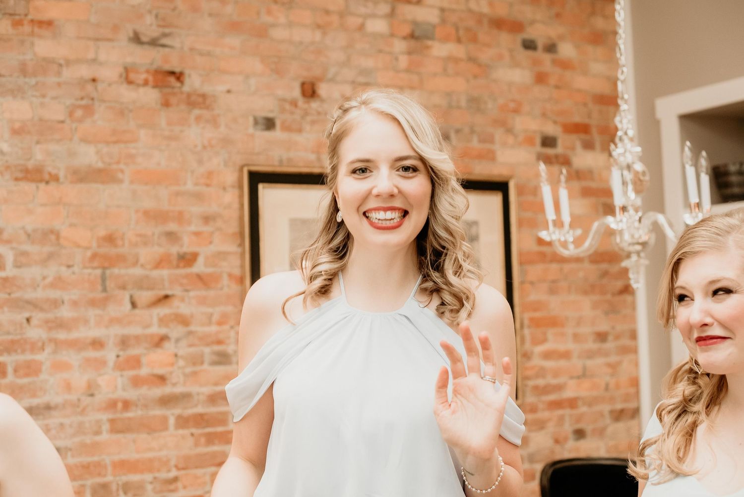Smiling blonde bridesmaid in gray off-shoulder dress waving, standing near brick wall with chandelier.