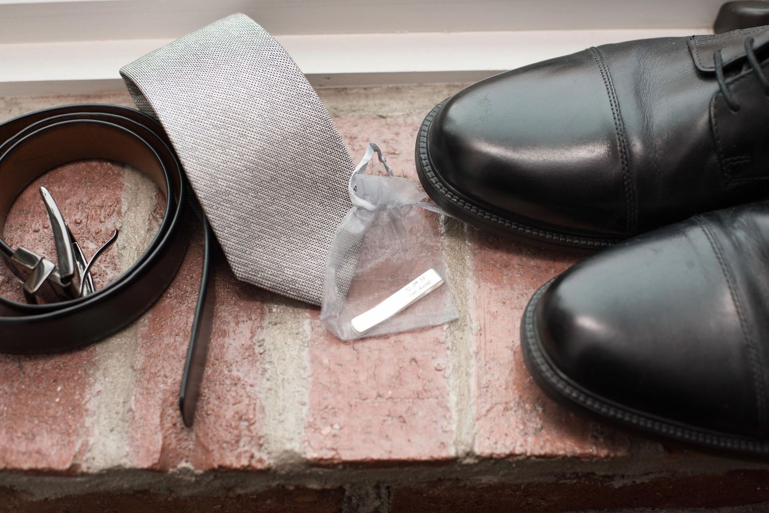 Black dress shoes, leather belt, gray tie, and cologne arranged on brick surface for a formal mens outfit flatlay.