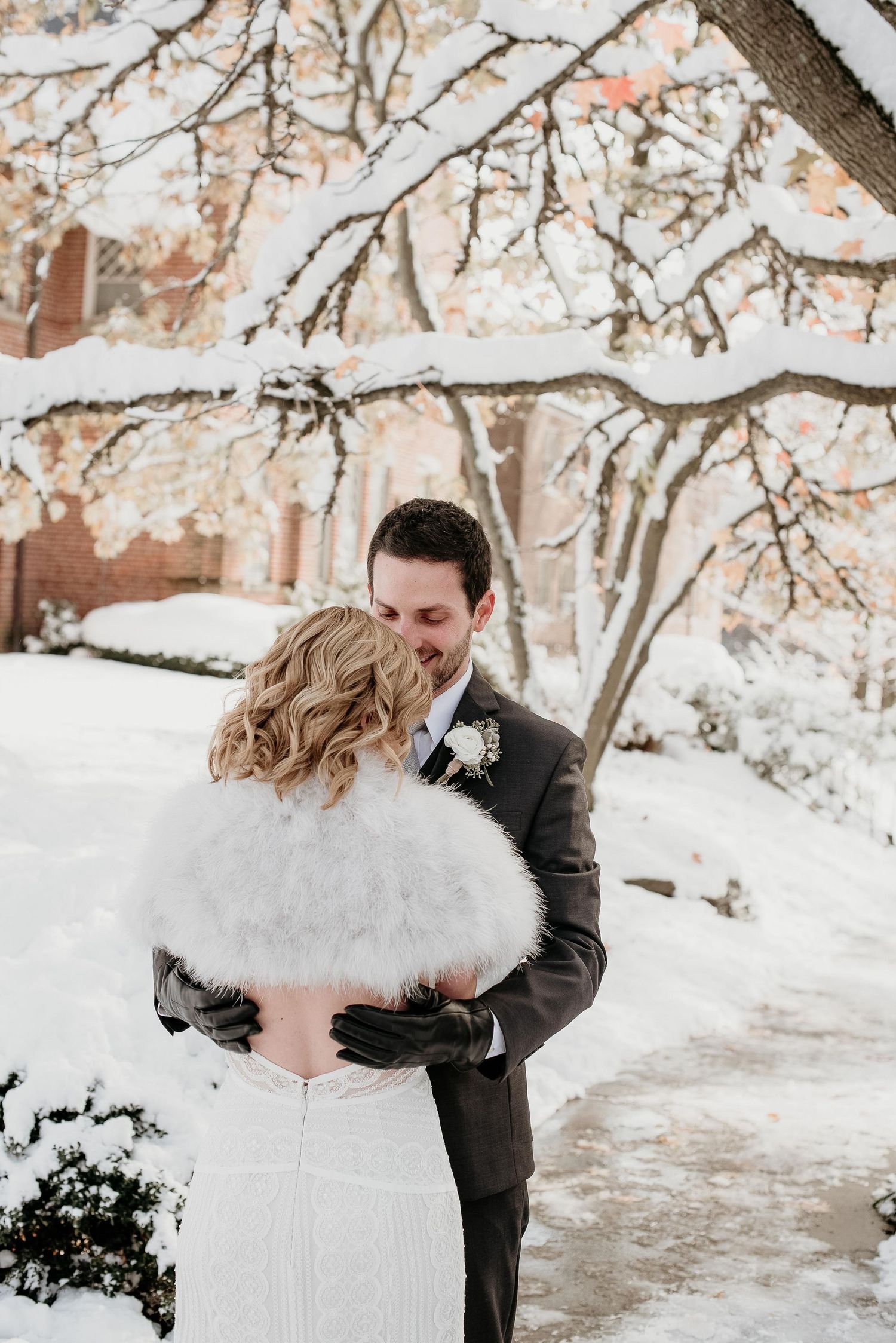 Couple embracing in snowy winter wedding scene, bride in white fur stole, groom in dark suit, snow-covered trees backdrop.